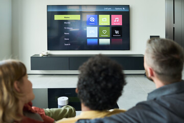 Rear view of diverse couple and child watching television screen in modern living room, with remote...