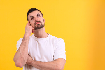 A young man is standing against a bright yellow background, looking up thoughtfully with a hand on...