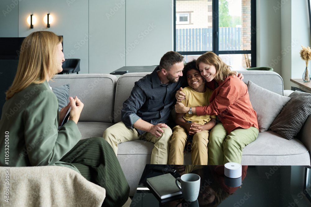 Wall mural Family gathered on living room sofa with two parents embracing their child, while another family member engages in conversation from an armchair, creating warm, intimate moment