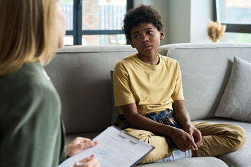 Young child sitting on couch interacting with therapist holding clipboard engaging in conversation...