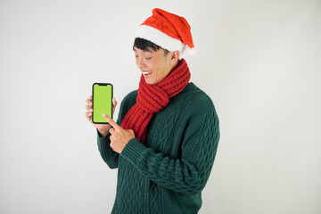 Young asian man wearing santa clause hat, red scarf and green long sleeved sweater is showing smartphone green screen, isolated over white background.