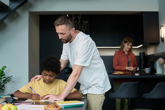 Father assisting young son with homework at kitchen table while mother is preparing meal in background, portraying a family moment of support and togetherness