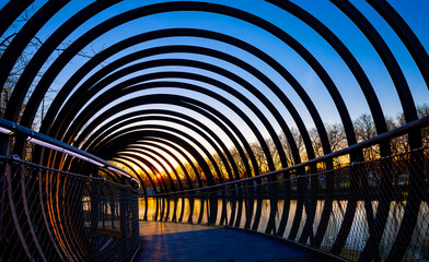 Naklejka premium Pedestrian bridge with steel loops spanning over a inland channel “Rhein-Herne-Kanal“ in Oberhausen Ruhr Basin, Germany. Colorful sunset with silhouettes of bare trees. Warm evening atmosphere.