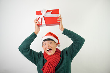 Young asian man wearing santa clause hat, red scarf and green long sleeved sweater with smiling face expression is carrying large gift box with ribbon, isolated over white background.