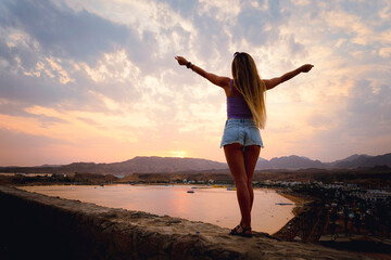 view from the back of the girl raising her hands up against the background of a beautiful dramatic sunset in a cloudy sky. Against the background of a coastal resort town.