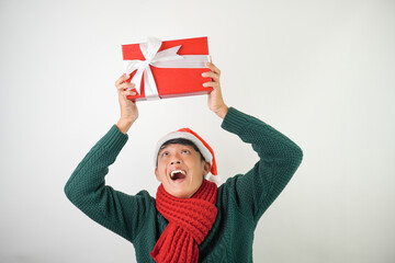 Young asian man wearing santa clause hat, red scarf and green long sleeved sweater with smiling face expression is carrying large gift box with ribbon, isolated over white background.