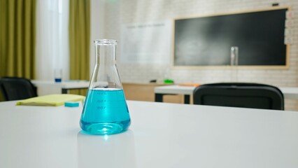 Empty school chemistry classroom with desks, chairs and chalkboard, workspace for teaching and studying. Glass bottle with blue liquid on table.