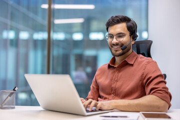 Smiling professional wearing headset working on laptop in modern office. Engaged in online communication, exemplifying technology's role in business. Reflects technology, and customer service.