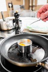 A close-up of a hand placing an egg inside a ring mold in a hot frying pan. The scene captures the cooking process in a professional kitchen, highlighting culinary preparation techniques.