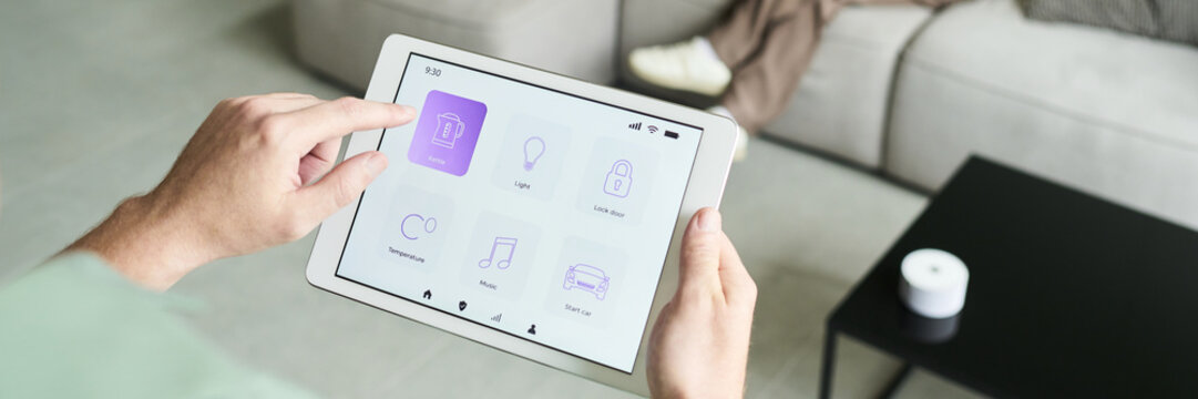 Person using tablet for controlling home automation system while seated on couch in modern living room. Displaying different icons for various home devices