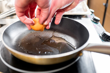 A chef's hand cracks an egg into a heated frying pan, showcasing the cooking process. The pan has a small amount of oil, with steam rising, indicating a professional kitchen setting.
