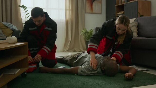 Emergency paramedics entering living room, using shining penlight into eyes of unconscious Black man with diabetes after severe hypoglycemia