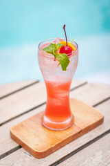 Singapore sling cocktails in glass garnished with slice of lemon, sprig of mint and cocktail cherry. The glass sits on a wooden tray