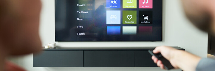 Two individuals seated on a couch are watching streaming media on a modern flat-screen TV mounted...