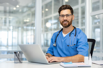 Confident doctor in blue scrubs smiling at camera while using laptop in bright office space....