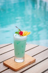 Blue hawaii in glass garnished with slice pineapples and cherry and mint leaves on wooden planks in front of a pool