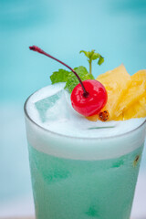 Close up view, Blue hawaii in glass garnished with slice pineapples and cherry and mint leaves on wooden planks in front of a pool