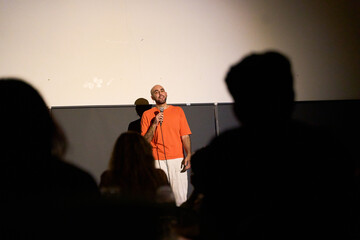 Male performer with shaved head delivering a monologue on stage in an alternative theater with...