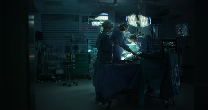 Diverse Surgical Team Working in a Darkened Operating Room Surrounded by Advanced Medical Equipment. Female Doctor Using a Scalpel and a Laparoscope while Performing a Complex Surgery
