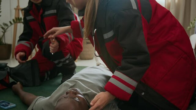 Female emergency paramedic using penlight to evaluate neurological response of unconscious Black man lying on floor after hypoglycemia, while her colleague preparing medical equipment nearby