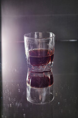 A glass tumbler filled with dark liquid resting on a reflective surface in a dimly lit setting