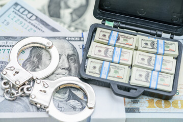 Financial crime, fraud, money laundering, corruption, or law enforcement : Bundles of US dollar 100 bills in a small case, alongside a pair of handcuffs placed over the currency.