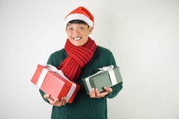 Young asian man wearing santa clause hat, red scarf and green long sleeved sweater with smiling face expression is carrying large gift box with ribbon, isolated over white background.