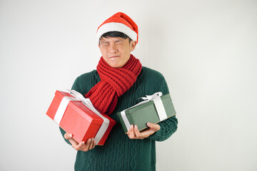 Young asian man wearing santa clause hat, red scarf and green long sleeved sweater with smiling face expression is carrying large gift box with ribbon, isolated over white background.