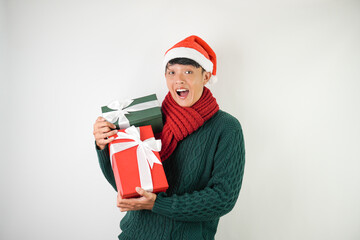 Young asian man wearing santa clause hat, red scarf and green long sleeved sweater with smiling face expression is carrying large gift box with ribbon, isolated over white background.