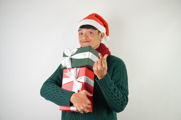 Young asian man wearing santa clause hat, red scarf and green long sleeved sweater with smiling face expression is carrying large gift box with ribbon, isolated over white background.