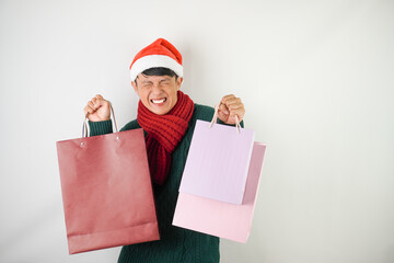 Young asian man wearing santa clause hat, red scarf and green long sleeved sweater with smiling face expression is carrying many shopping bags, isolated over white background.