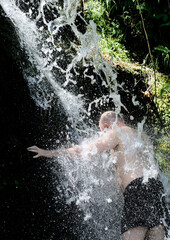 A man stands under the clear water of a waterfall and the numerous splashes hide him behind the water.