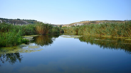 A calm river flows through a landscape bordered by thick reeds, chalk mountains, under a bright blue sky.