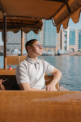 Young arab male tourist taking a ride on abra boat, traditional water transport in the UAE. Lebanese man ejoying city tour sitting in a boat with his earphones and closed eyes listening to music