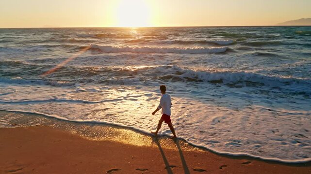 Young man with white shirt walking alone along the beach enjoying the beautiful sunset. waves coming to the shore. Sun is setting. Warm dusk sky. Golden hour. Summer holidays vibes.