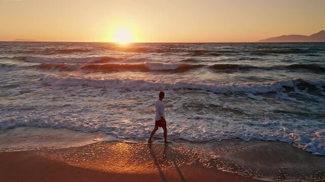 Young man with white shirt walking alone along the beach enjoying the beautiful sunset. waves coming to the shore. Sun is setting. Warm dusk sky. Golden hour. Summer holidays vibes.