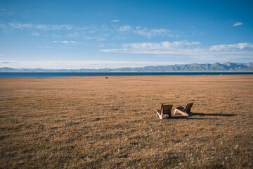 Relaxing view of wooden chairs near the tranquil waters of Song-Kol Lake, surrounded by vast grasslands and mountains, Kyrgyzstan, Central Asia