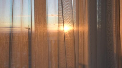Sunlight streaming through sheer curtains creates warm, golden glow during a stunning sunrise over city skyline, filling the home interior with peaceful and cozy ambiance. Window view from apartments