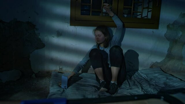 Full low-key shot of young Caucasian captive lady sitting alone in dark basement handcuffed to window grate, she removing duct tape from mouth, taking phone and calling 911, human abduction