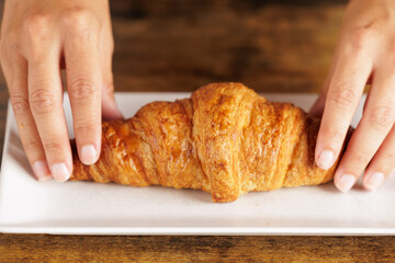 Close-up of a woman's hands holding a plate with a freshly baked croissant. Shop assistant serves a handmade croissant in the coffee. Sweet or savory breakfasts or snacks.