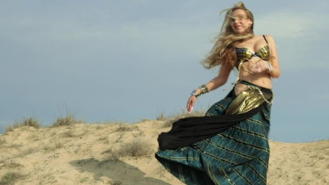 Dancer Weare Gold and Blue Bedlah is Moving Gracefully in Sand Dunes. Girl Practicing Belly Dance Routine Outdoors