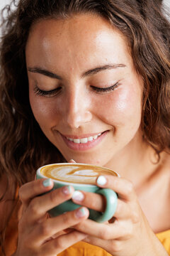 Portrait of a young woman as she smiles and enjoys the aroma of the specialty coffee she has in her hands. Coffee with a drawing of a heart. Concept of tranquility and calm, tasting a good coffee.