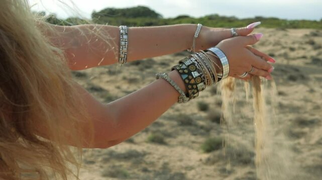 Blond woman wearing bracelets lets sand fall through her hands against stunning desert landscape, embodying freedom and beauty in serene, sunlit environment