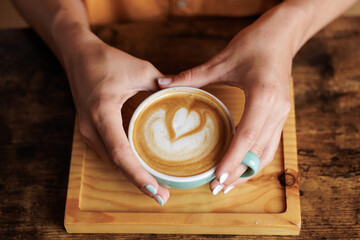 Woman's hands holding a blue cup of specialty coffee with a heart drawn on it. Woman at break time from work or student relaxing after exams.