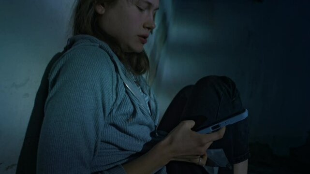 Side tilt handheld shot of young scared Caucasian lady being held hostage in dark basement, removing duct tape from mouth and quickly calling rescue service using smartphone