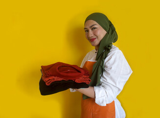 Side View Of Smiling Asian Muslim Woman Holding sSack Of Folded Clothes On Yellow Background