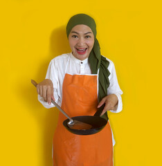 Young Asian Muslim Woman Housewife Wearing Kitchen Apron Cooking And Holding Pan And Spatula Isolated On Yellow Background