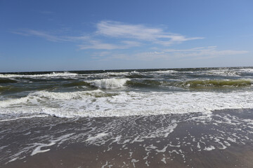 Scenic view of ocean waves crashing onto the sandy shore, with a clear blue sky and scattered clouds in the background