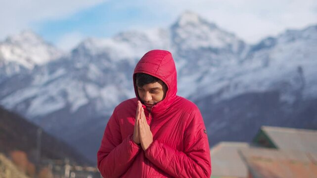 4K Portrait shot of an Indian man wearing jacket rubbing his hands in front of snowy Himalayan mountain peaks during the winter season at Keylong in Lahaul, Himachal Pradesh, India. Cold during winter