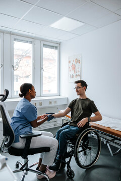 Female medical professional doing hand stretch of teenage boy sitting on wheelchair in examination room